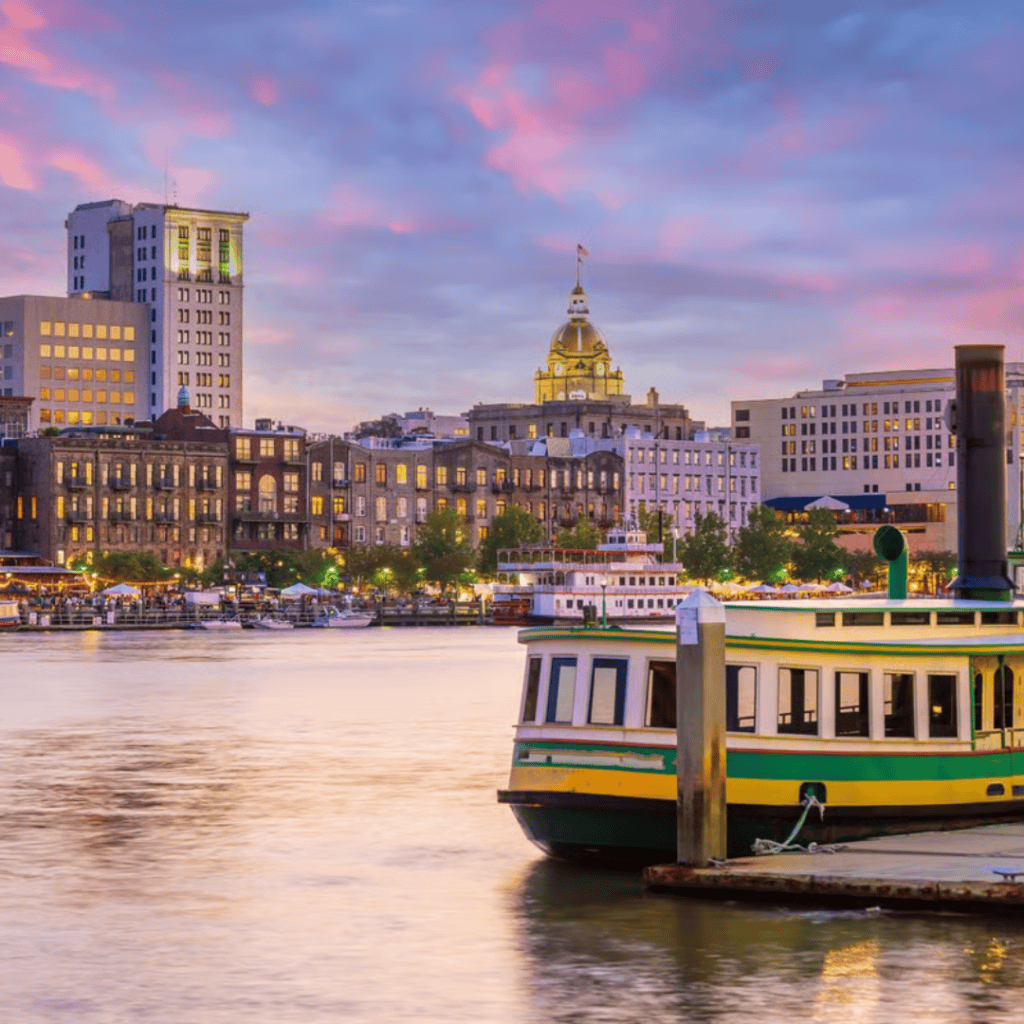 Savannah, Georgia waterfront at sunset with historic riverfront buildings, glowing city lights, a riverboat docked along the harbor, and the gold dome of City Hall under a colorful evening sky.